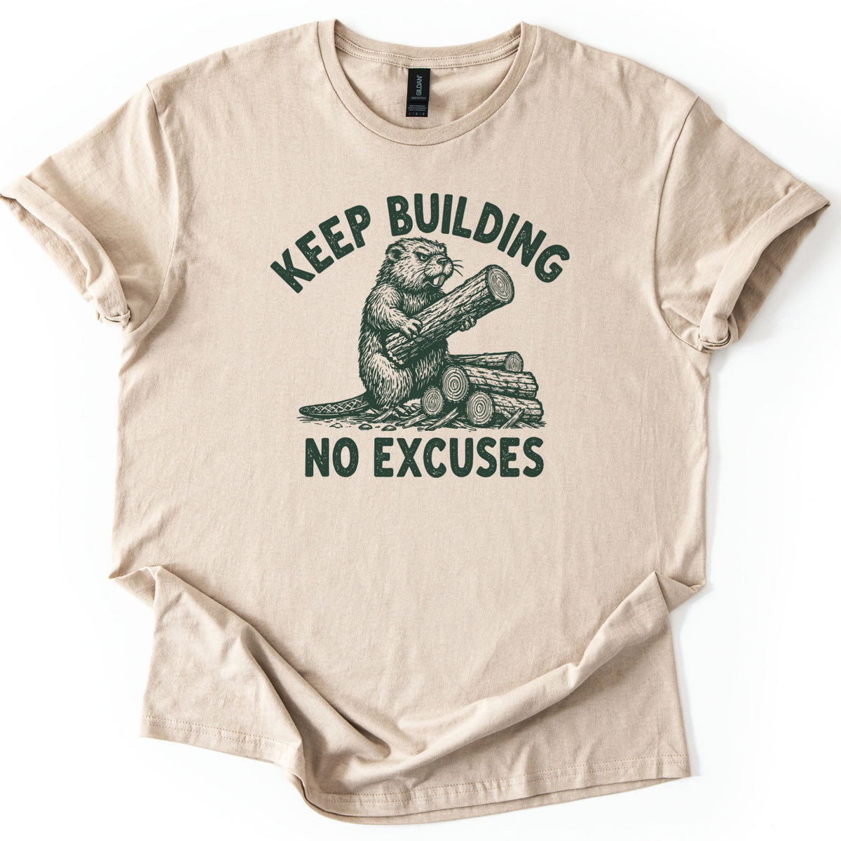Keep Building Beaver Comfort Colors T-shirt in Sand, laid flat. Dark green sketch-style artwork of a beaver hugging a log while sitting on a pile of cut logs, with Keep Building arched above and No Excuses below. Sleeves are rolled on a white background.