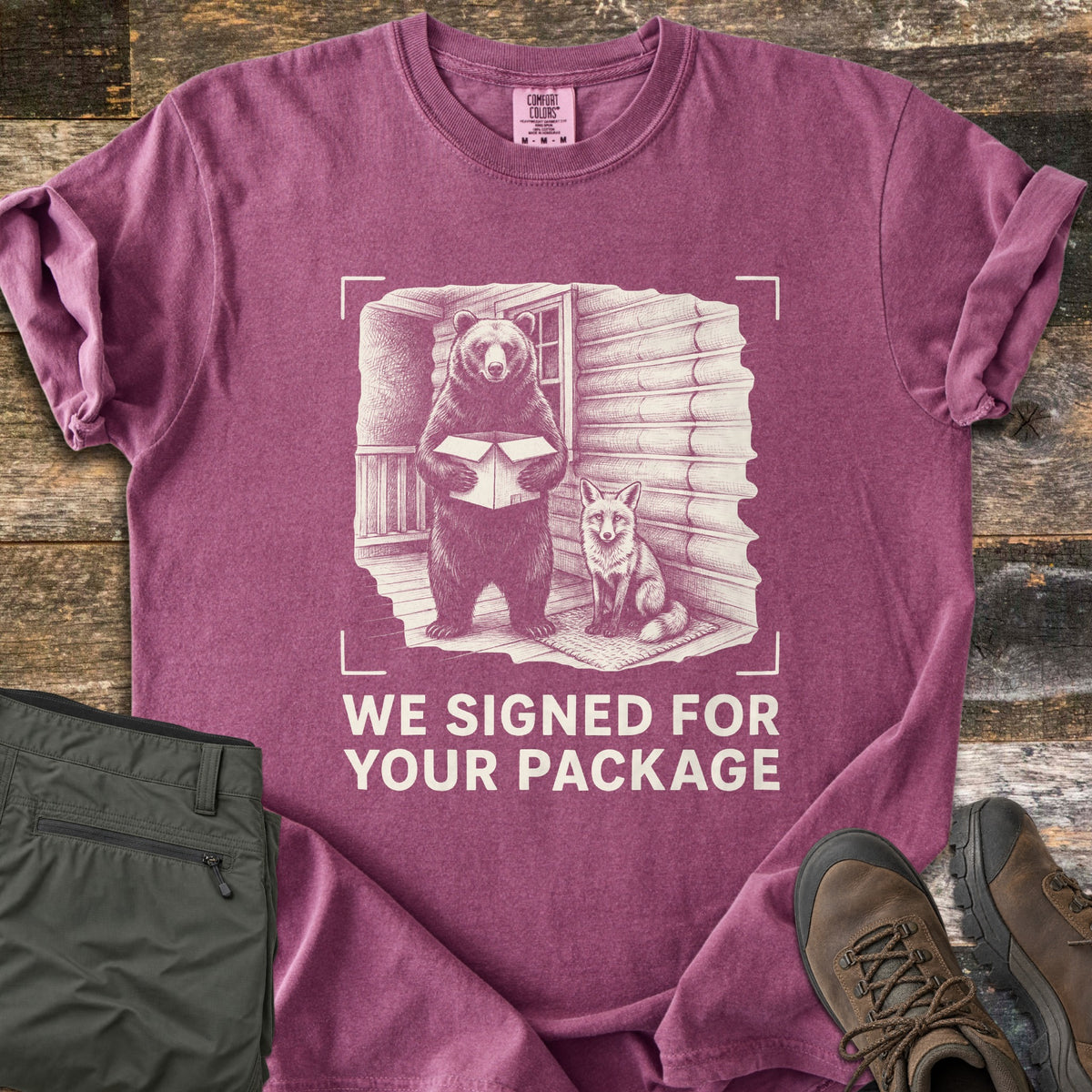 We Signed For Your Package Comfort Colors T-shirt in Berry, laid flat. A cream-ink sketch shows a bear holding an open box beside a seated fox on a log cabin porch, framed by camera corner marks above the words We Signed For Your Package. Olive pants and brown hiking boots sit on wood planks.