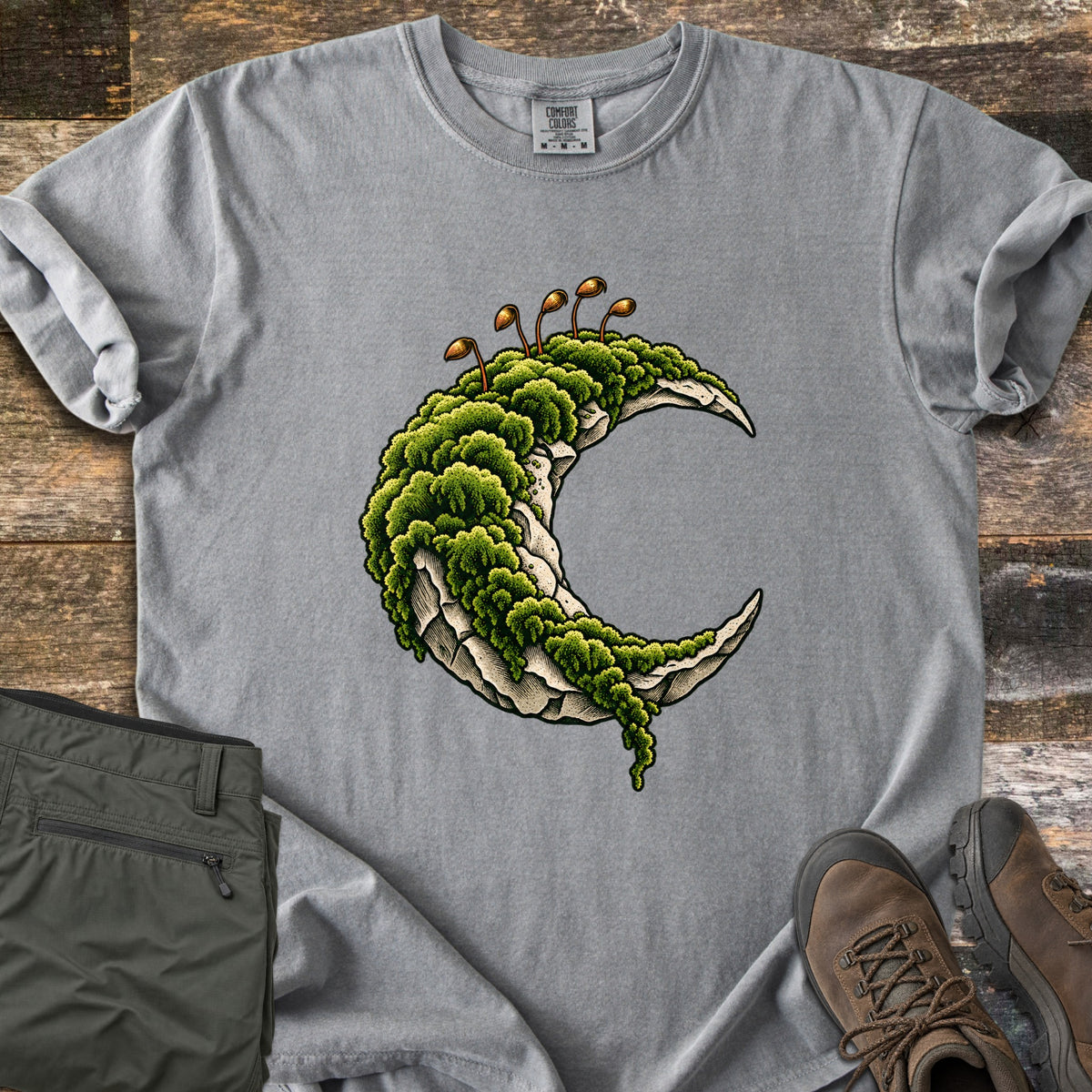 Moss Moon Comfort Colors T-shirt in Grey, laid flat. A pale stone crescent moon draped in dense green moss, with thin brown spore stalks on top. Sleeves rolled, on rustic wood planks next to folded olive hiking pants and a pair of brown leather hiking boots.