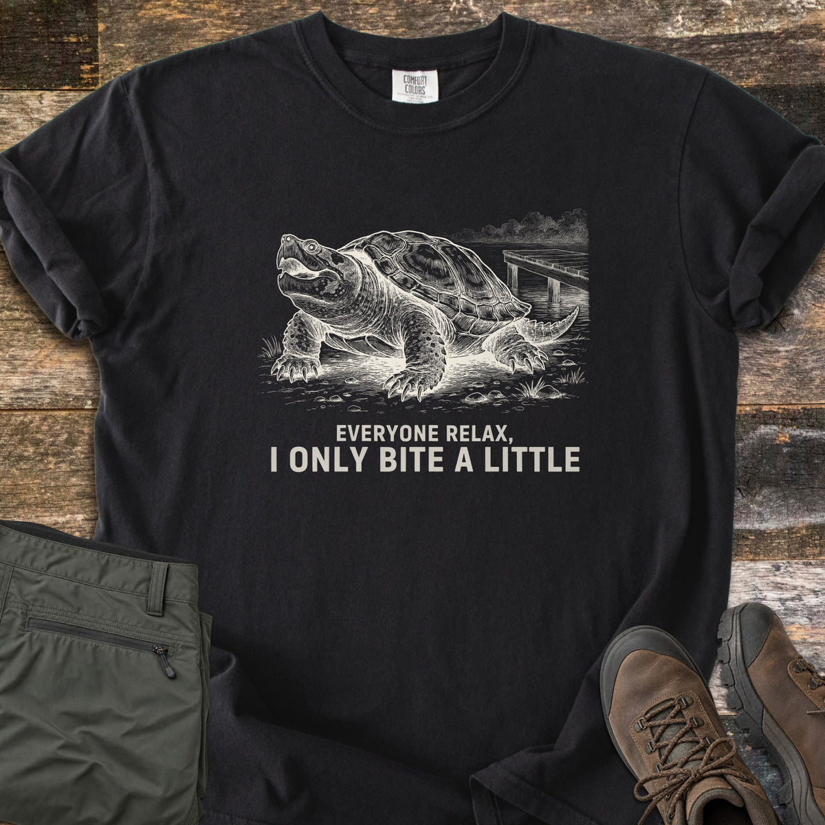 Everyone Relax I Only Bite A Little Comfort Colors T-shirt in Black, laid flat. A white sketch of a snapping turtle with open mouth on a lakeshore by a wooden dock, above the words Everyone relax, I only bite a little. Olive hiking pants and brown hiking boots sit on a weathered wood floor.