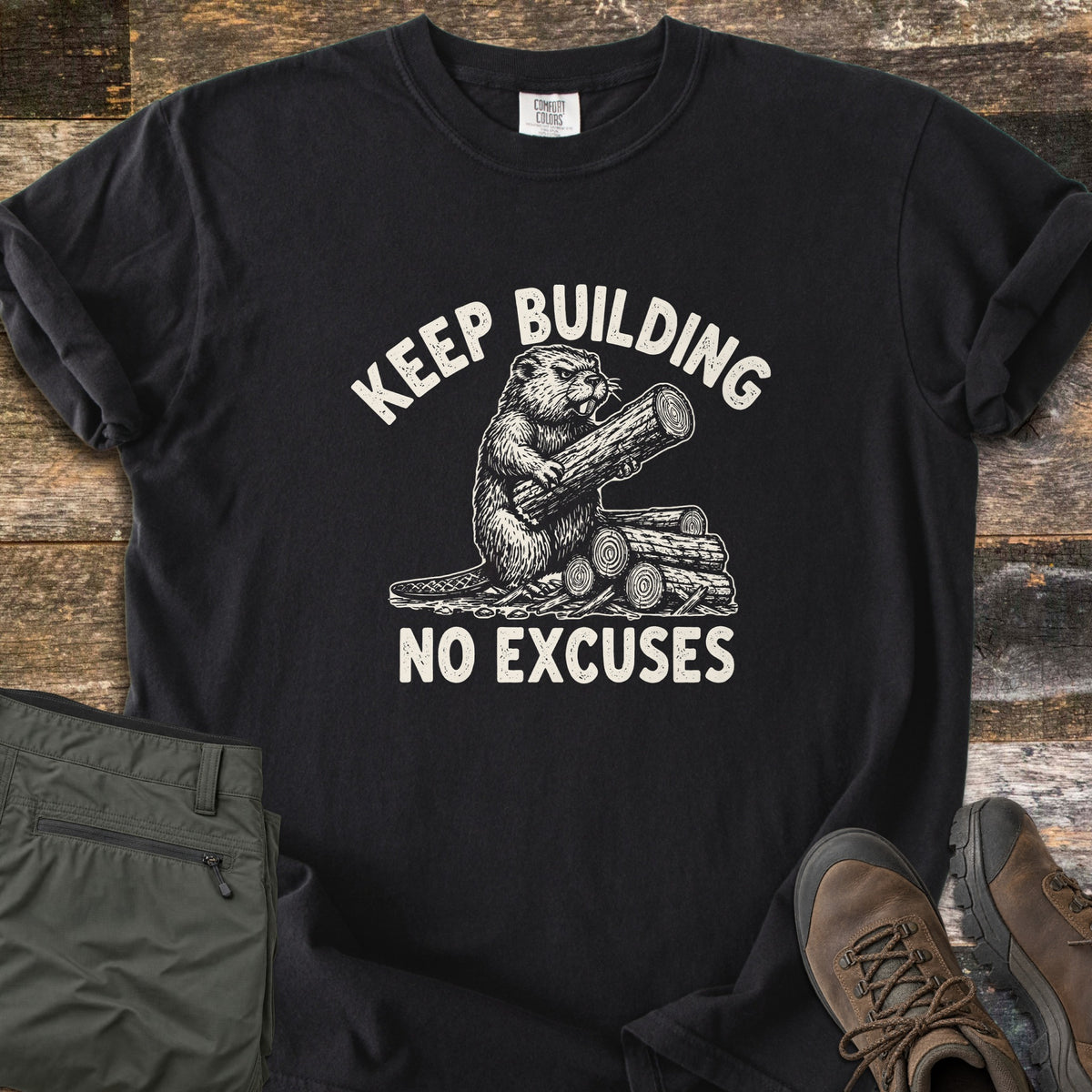 Keep Building Beaver Comfort Colors T-shirt in Black, laid flat. A white vintage-style print of a beaver hugging a log beside a pile of cut logs, with the words KEEP BUILDING arched above and NO EXCUSES below. Shown on weathered wood with olive pants and brown hiking boots.
