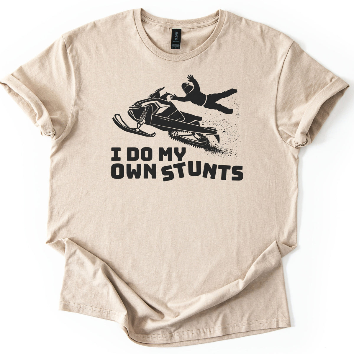 Snow Mobile Stunts Comfort Colors T-shirt in Sand, laid flat. A black silhouette of a snowmobile flying through the air, its rider thrown off behind it in a spray of snow, above the words I Do My Own Stunts. The shirt lies on a white background with rolled sleeves.