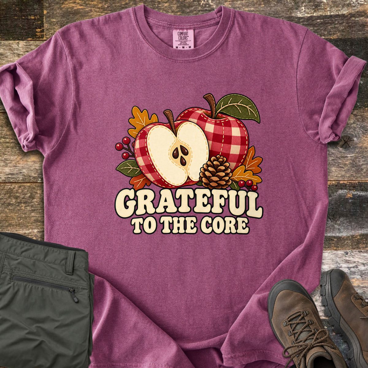 Grateful to the Core Comfort Colors T-shirt in Berry, laid flat. A red gingham apple and an apple half with seeds sit with a pinecone, autumn leaves and berries above the words Grateful to the Core. Sleeves rolled, it lies on weathered wood by olive pants and brown boots.