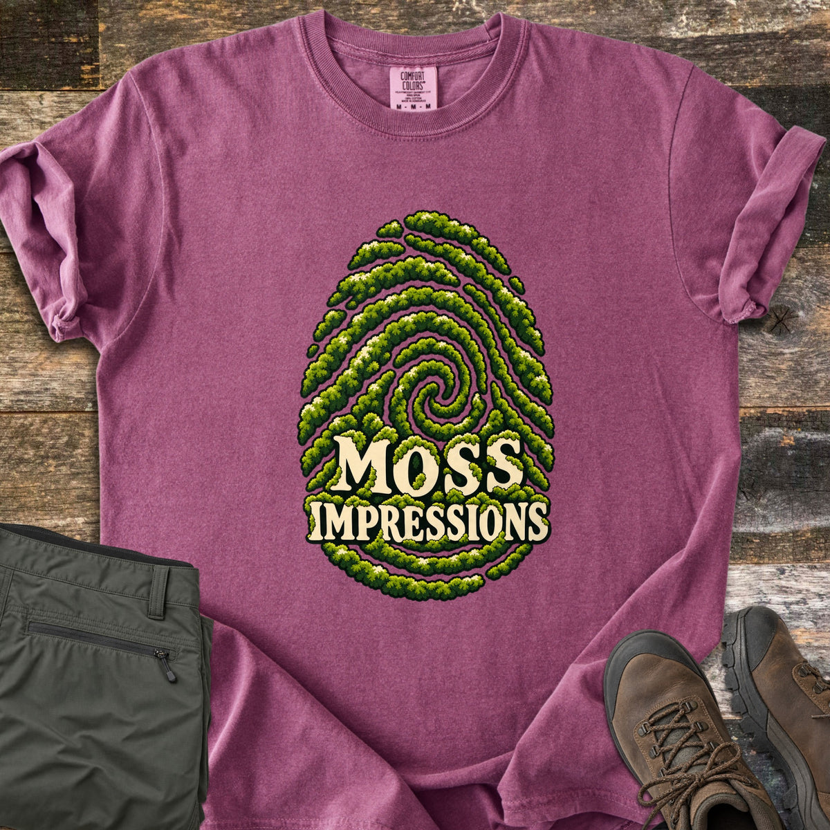 Moss Impressions Comfort Colors T-shirt in Berry, laid flat. A fingerprint made of green moss ridges with the words Moss Impressions in cream letters. The shirt lies on weathered wood with folded olive hiking pants and brown hiking boots.