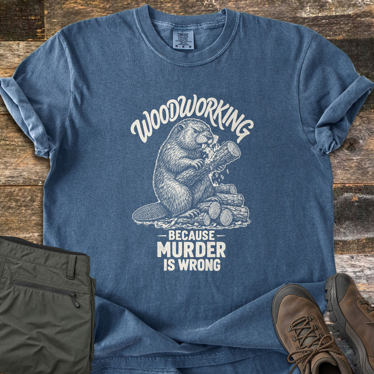 Woodworking Because Murder Is Wrong Comfort Colors T-shirt in Denim, laid flat. A cream sketch of a beaver gnawing a log on a woodpile, under the arched words Woodworking and above Because Murder Is Wrong. Olive cargo pants and brown hiking boots sit beside it on weathered wood planks.