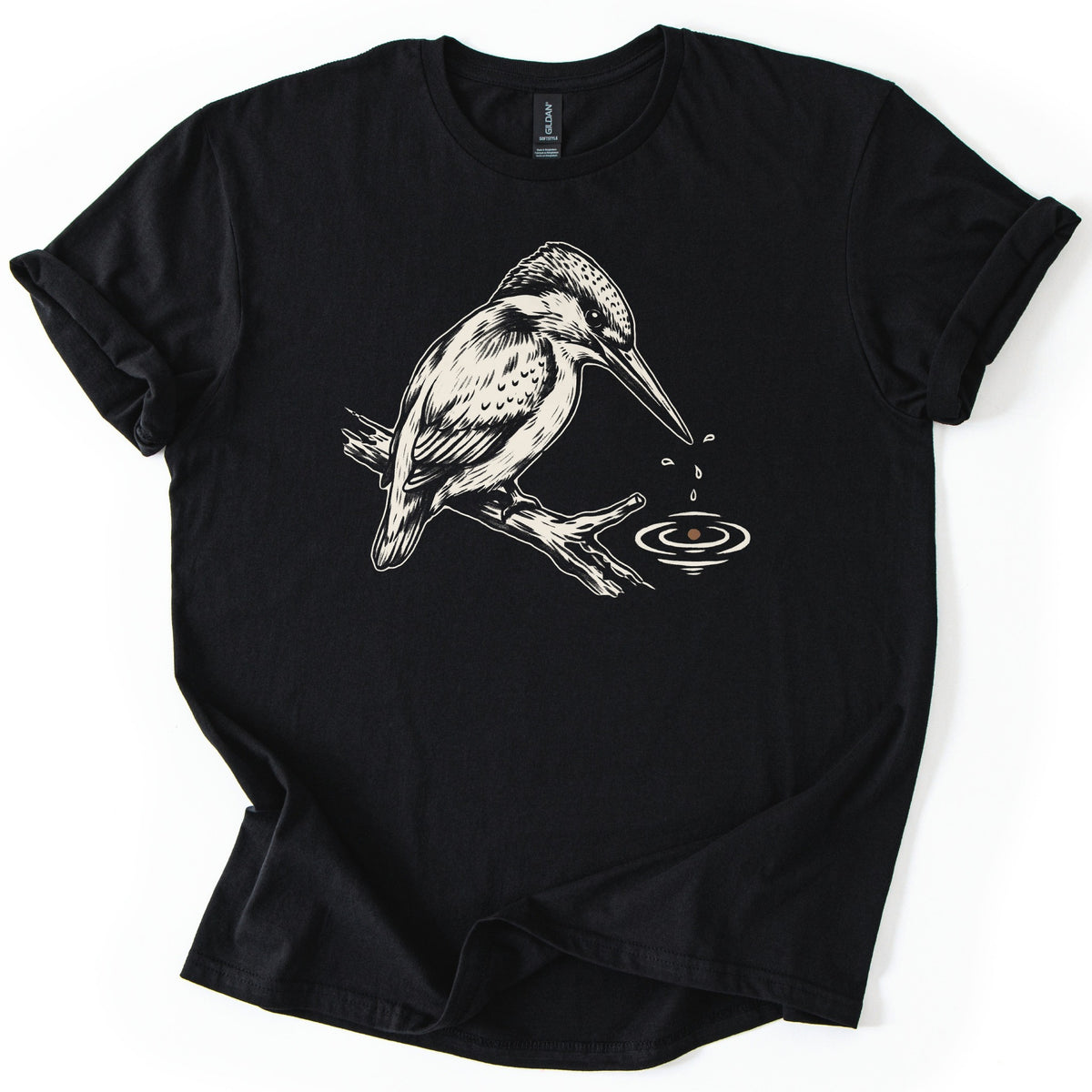 Kingfisher Comfort Colors T-shirt in Black, laid flat. A cream-colored drawing of a kingfisher perched on a branch, with droplets falling from its beak into rippling water below. The sleeves are cuffed and the shirt lies on a white background.