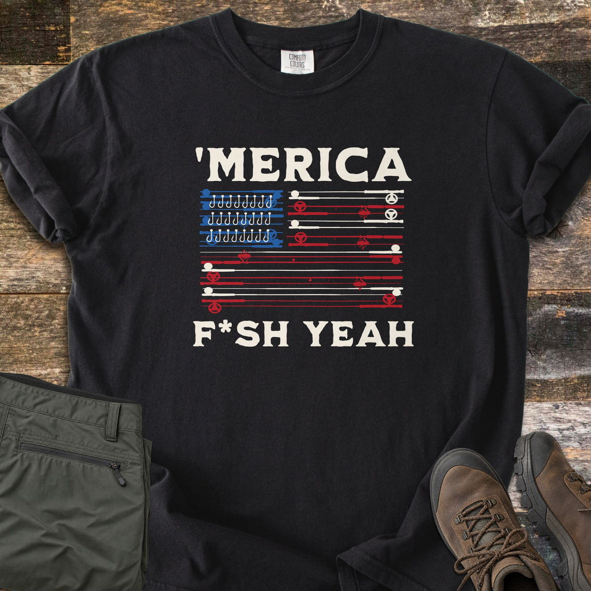 Fish America Fish Yeah Comfort Colors T-shirt in Black, laid flat. White text reads 'MERICA above an American flag made of red and white fishing rods, with rows of hooks in the blue corner, then F*SH YEAH. Folded olive pants and brown hiking boots sit on weathered wood.