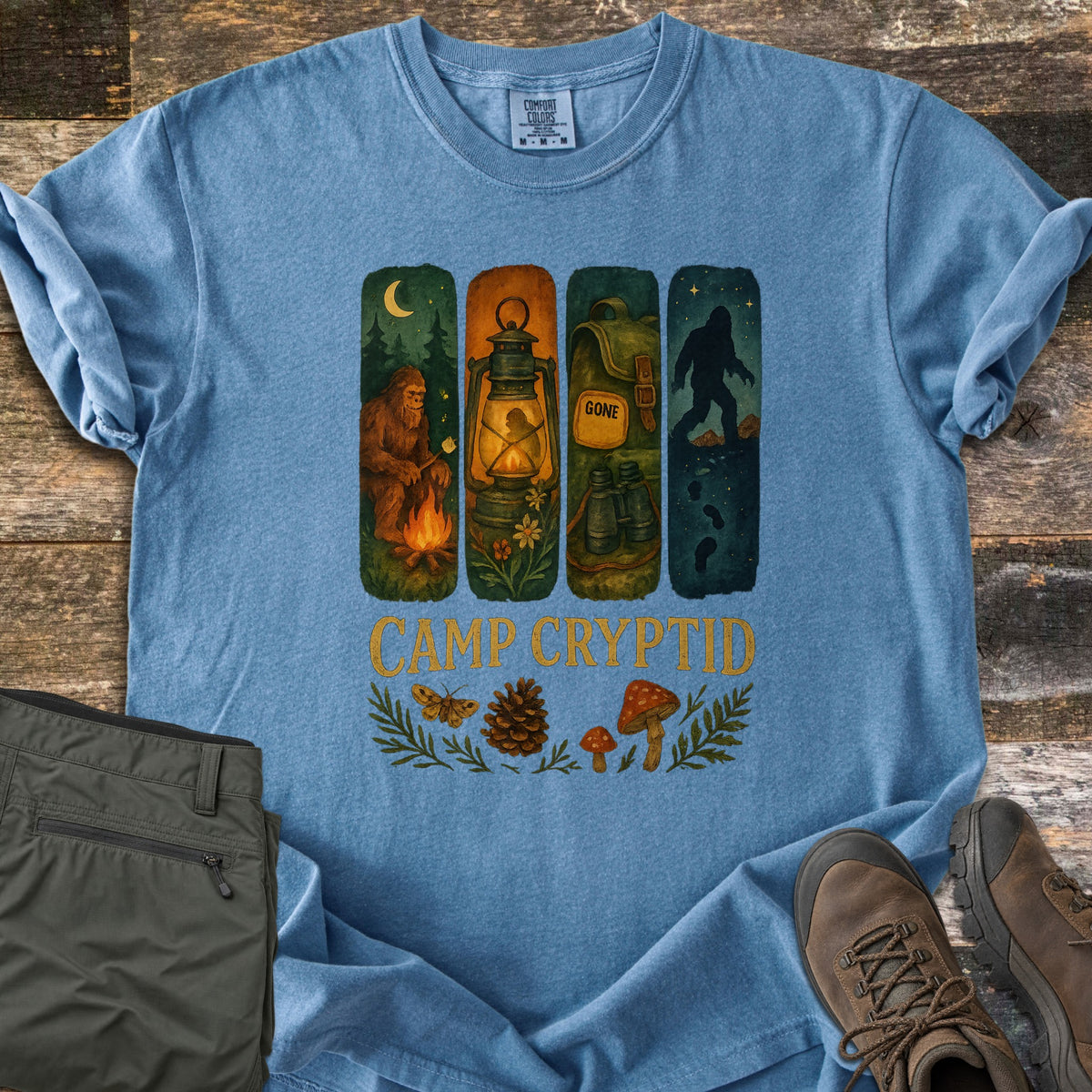 Camp Cryptid Big Foot Comfort Colors T-shirt in Blue Jean, laid flat. Four painted panels show Bigfoot at a campfire, a lantern, a backpack marked GONE with binoculars, and Bigfoot under stars, above CAMP CRYPTID, a moth, pinecone and mushrooms. Olive pants and hiking boots lie on wood.
