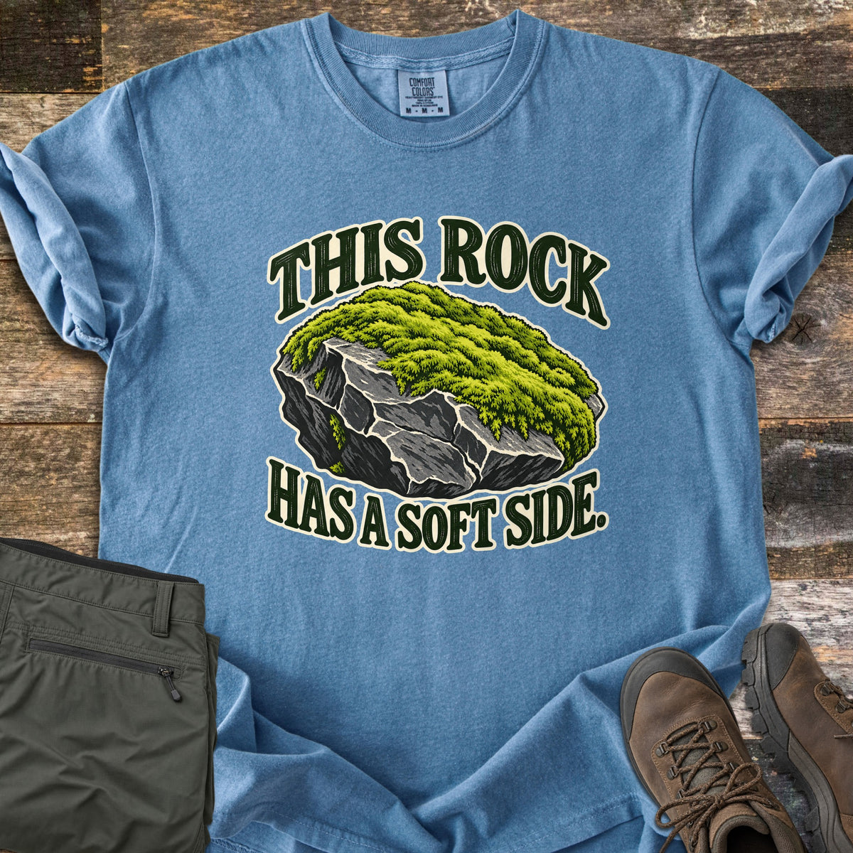 This Rock Has a Soft Side Comfort Colors T-shirt in Blue Jean, laid flat. An illustrated gray boulder topped with thick green moss sits between the words This Rock above and Has a Soft Side. below. Olive hiking pants and brown hiking boots lie beside it on weathered wood planks.