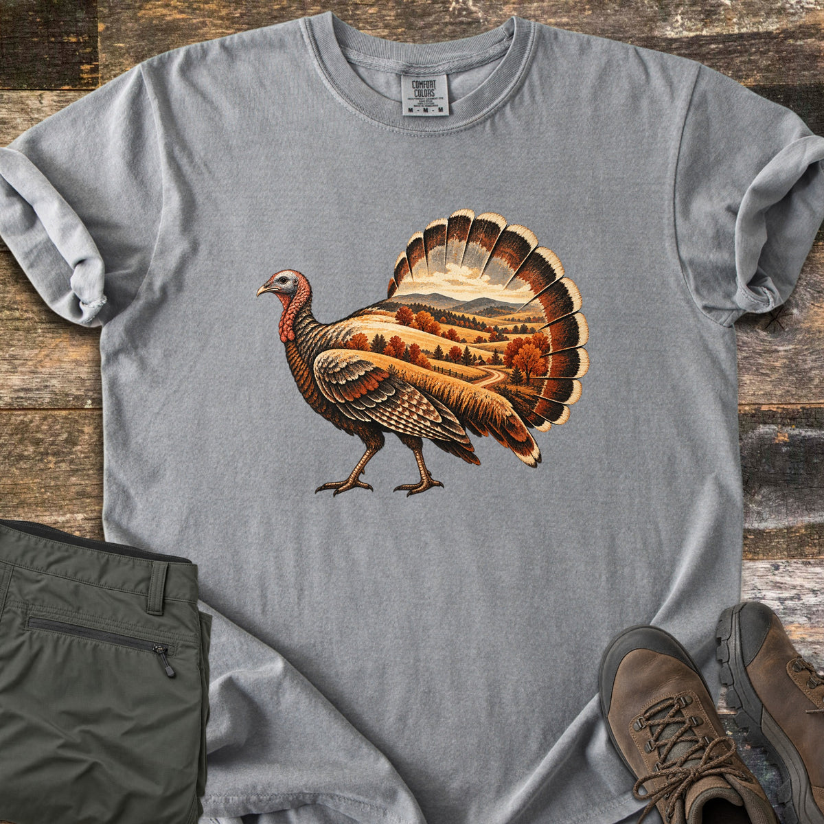 Turkey Harvest Hills Comfort Colors T-shirt in Grey, laid flat. A wild turkey in profile, its fanned tail and body showing fall hills, orange trees and a winding road under distant mountains. Sleeves rolled, on weathered wood planks with folded olive pants and brown boots.