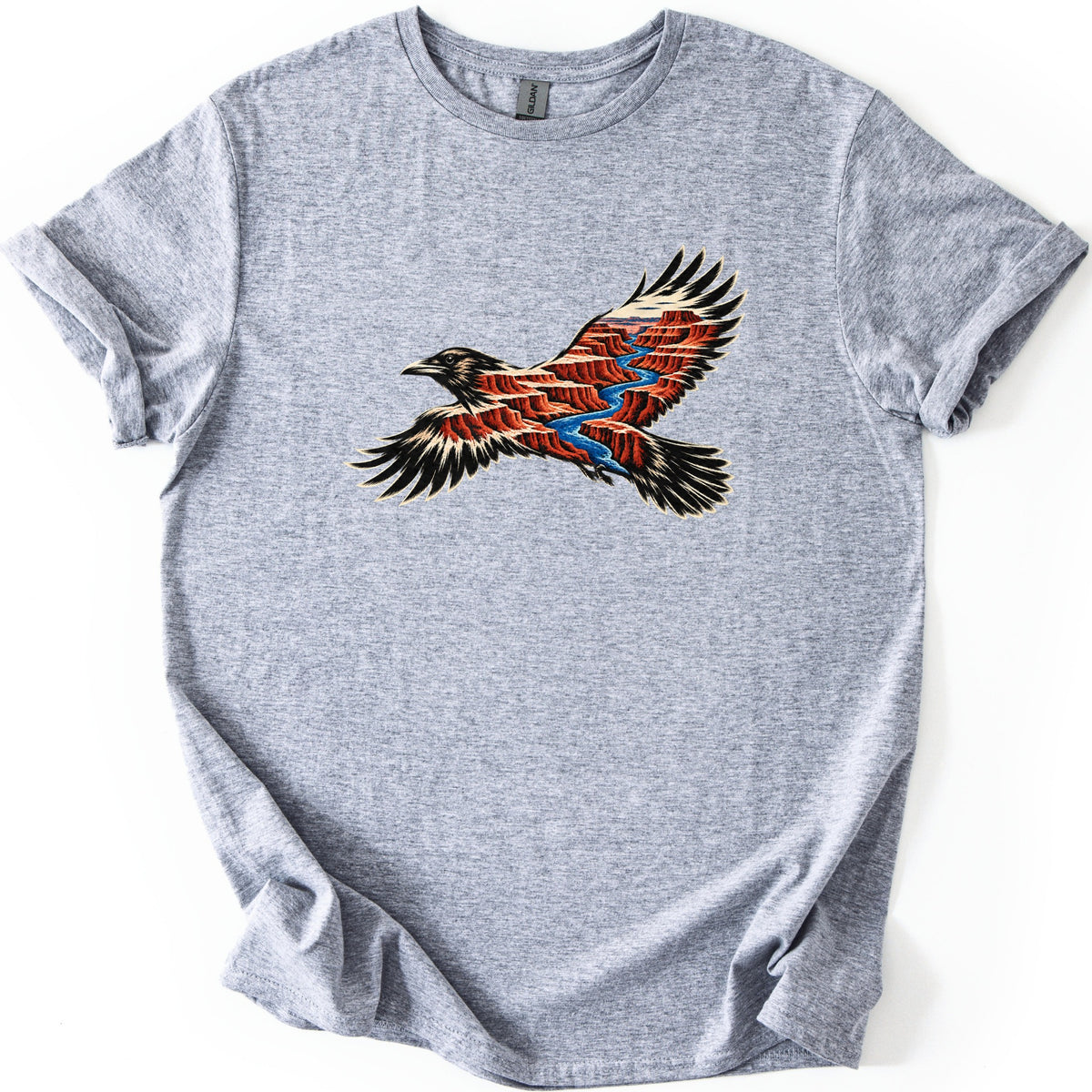 Red Canyon Raven Comfort Colors T-shirt in Sport Grey, laid flat. A black raven in flight, its body and wings filled with red canyon cliffs and a winding blue river. No words are printed. The shirt lies on a white surface with rolled sleeves and a loosely folded hem.