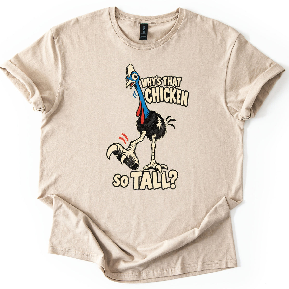 Why's That Chicken So Tall Comfort Colors T-shirt in Sand, laid flat. A cartoon cassowary with a blue neck, red wattles and long scaly legs kicks up one clawed foot next to cream text reading Why's That Chicken So Tall? The shirt lies on white, its hem loosely folded.