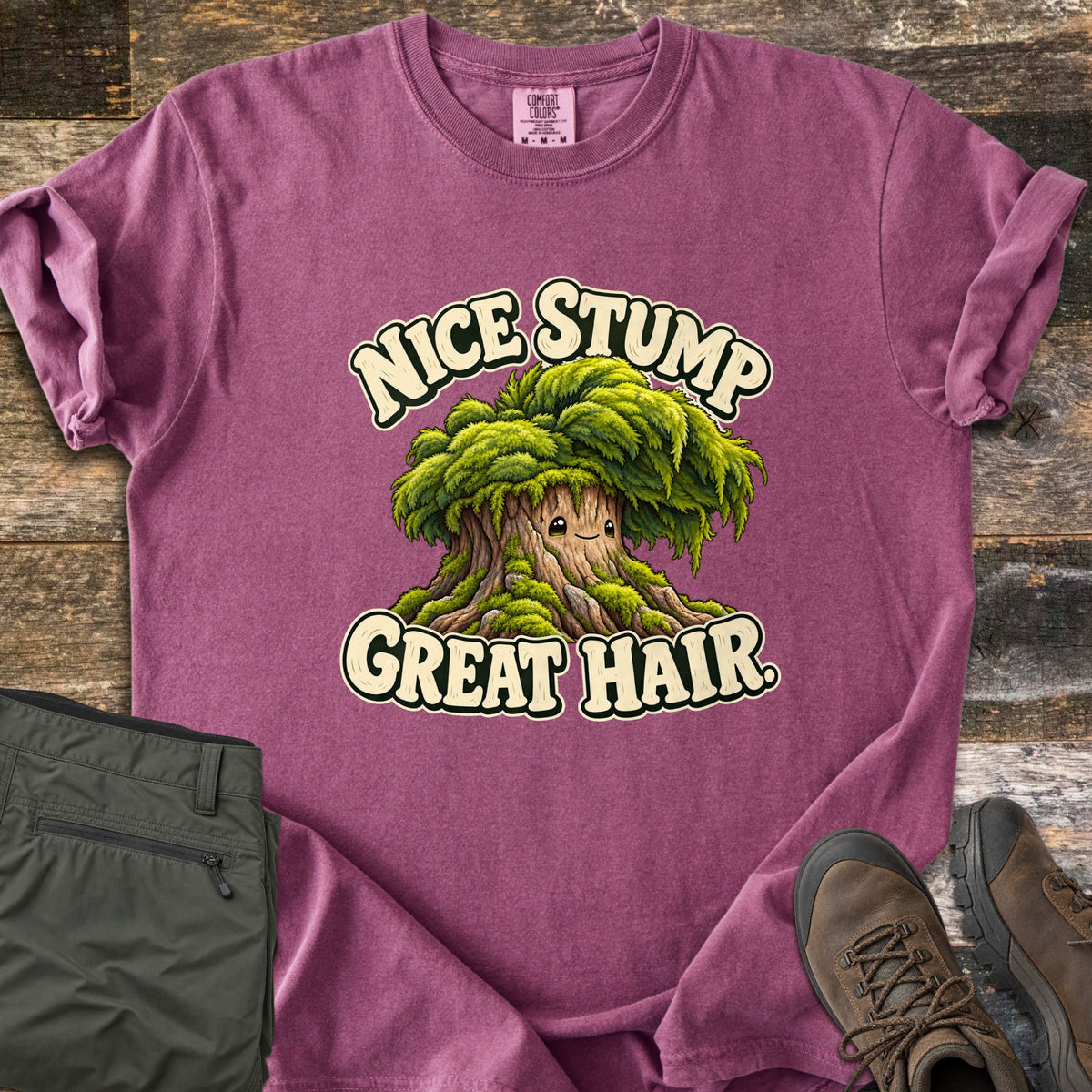 Nice Stump Great Hair Comfort Colors T-shirt in Berry, laid flat. A smiling cartoon tree stump with a shaggy mop of green moss hair sits between the words Nice Stump Great Hair. Sleeves rolled, on weathered wood with folded olive hiking pants and brown hiking boots.