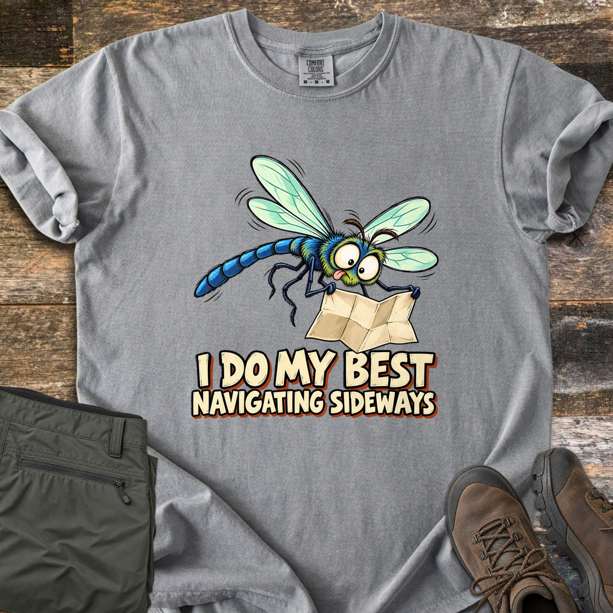 I Do My Best Navigating Sideways Comfort Colors T-shirt in Grey, laid flat. A wide-eyed cartoon dragonfly with its tongue out holds a folded paper map above the words I Do My Best Navigating Sideways. Olive pants and brown hiking boots lie beside it on weathered wood planks.