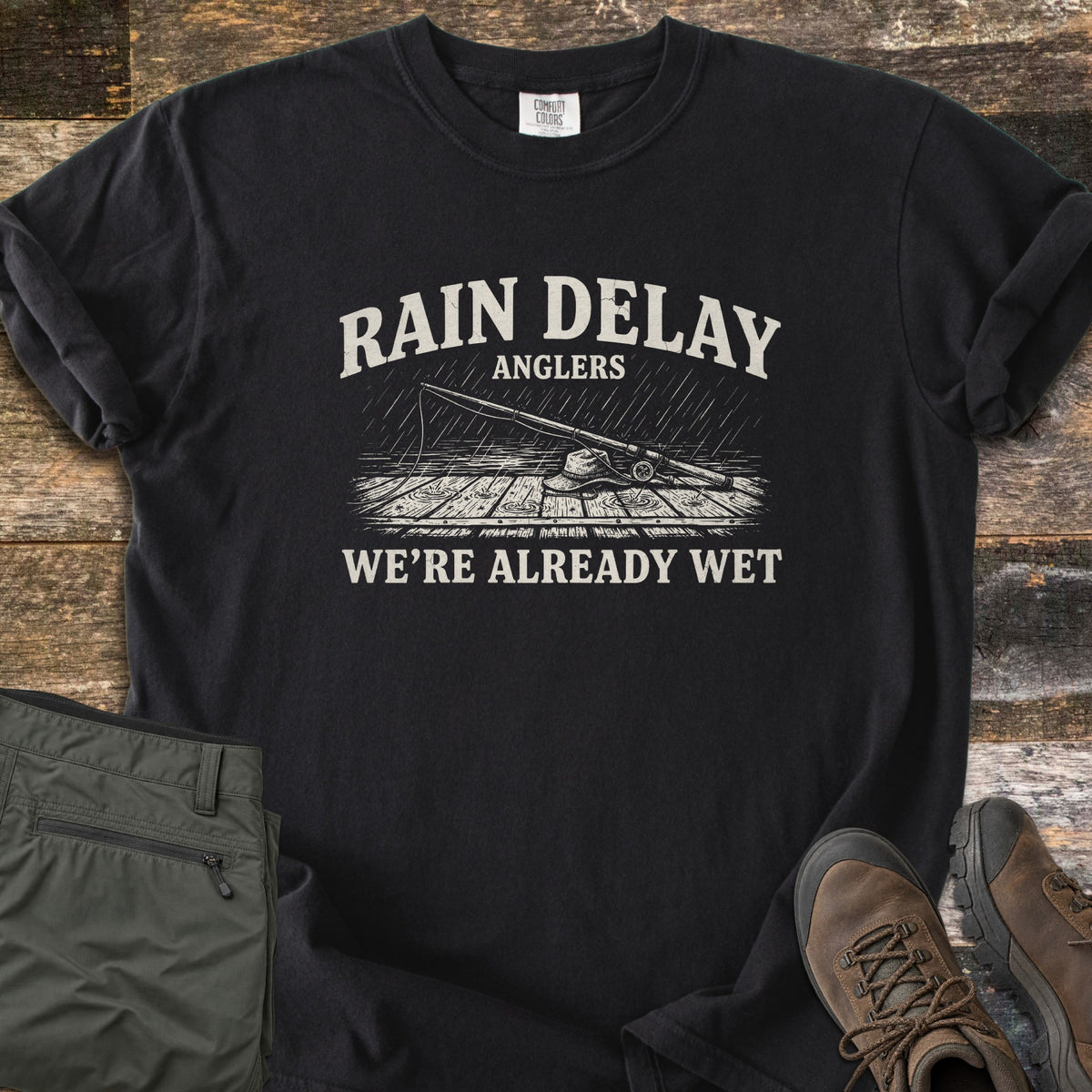 Rain Delay Anglers Comfort Colors T-shirt in Black, laid flat. Cream print of a fishing rod and bucket hat lying on a wet dock in the rain, with Rain Delay Anglers above and We're Already Wet below. On weathered wood planks next to olive hiking pants and brown boots.