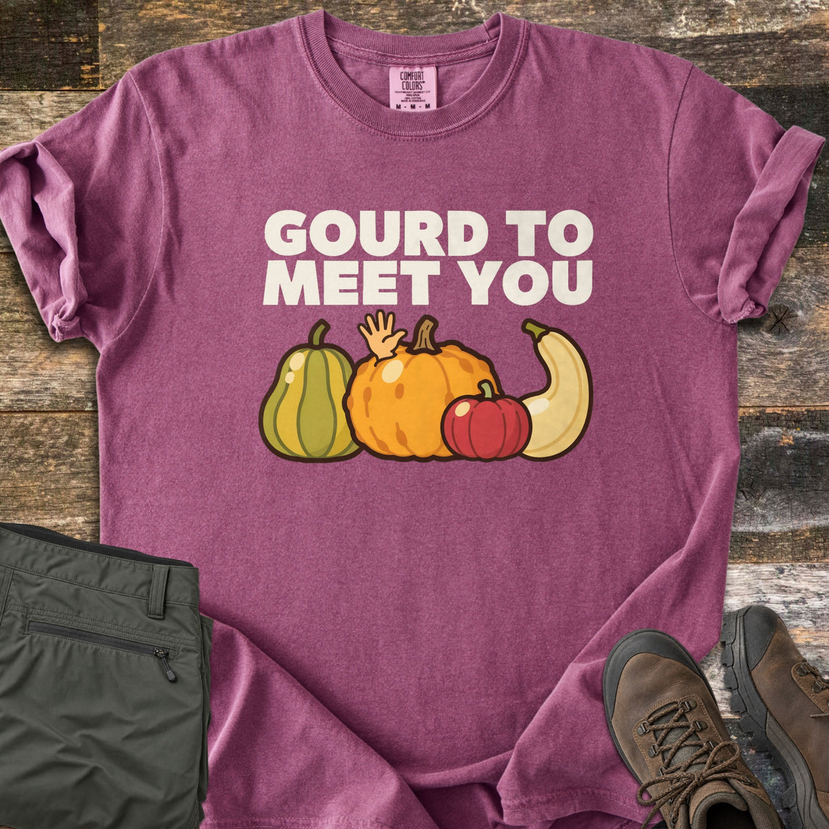 Gourd To Meet You Comfort Colors T-shirt in Berry, laid flat. Off-white words Gourd To Meet You above cartoon gourds: a green striped squash, an orange pumpkin with a waving hand, a red pumpkin and a cream squash. Olive pants and brown hiking boots sit on wood planks.