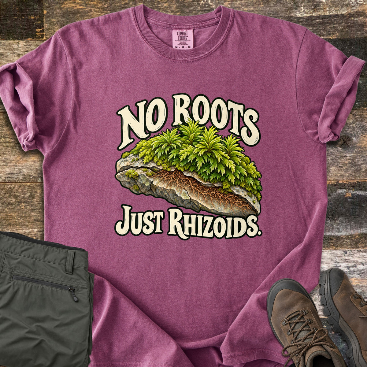 No Roots Just Rhizoids Comfort Colors T-shirt in Berry, laid flat. Green moss sprouts from a cracked rock with brown rhizoids threading underneath, between the words No Roots Just Rhizoids. On rustic wood beside folded olive hiking pants and brown hiking boots.
