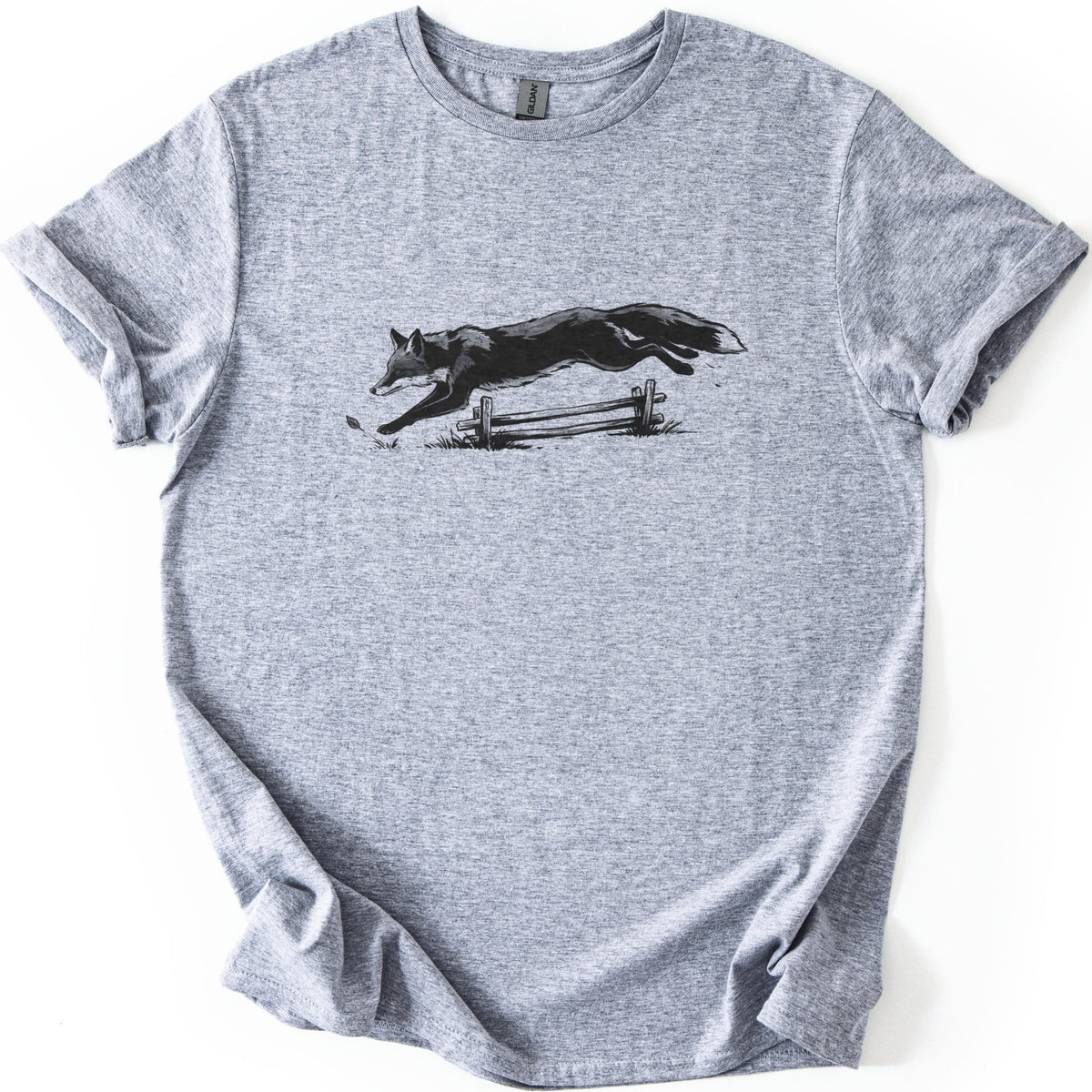 Leaping Fox Comfort Colors T-shirt in Sport Grey, laid flat. A black and gray sketch of a fox leaping over a low wooden rail fence, with grass tufts and a falling leaf below. The shirt has cuffed sleeves and lies on a white background.