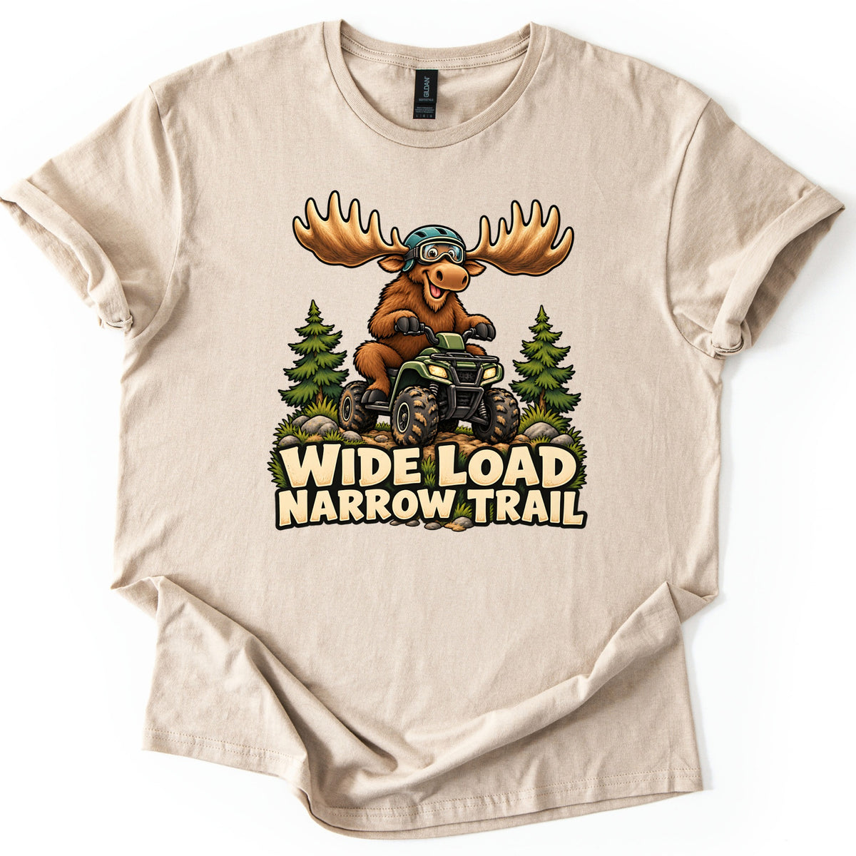 Wide Load Narrow Trail Comfort Colors T-shirt in Sand, laid flat. A cartoon moose with huge antlers, a helmet and goggles rides a green ATV on a dirt trail between pine trees above the words Wide Load Narrow Trail. The shirt lies on a white background with cuffed sleeves.