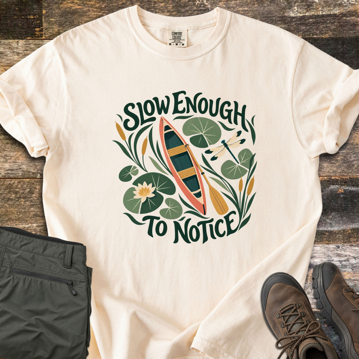 Slow Enough To Notice Comfort Colors T-shirt in Ivory, laid flat. A coral canoe with a yellow paddle floats among lily pads, a water lily, cattails and a dragonfly between the words Slow Enough To Notice. Folded green hiking pants and brown hiking boots sit on weathered wood.