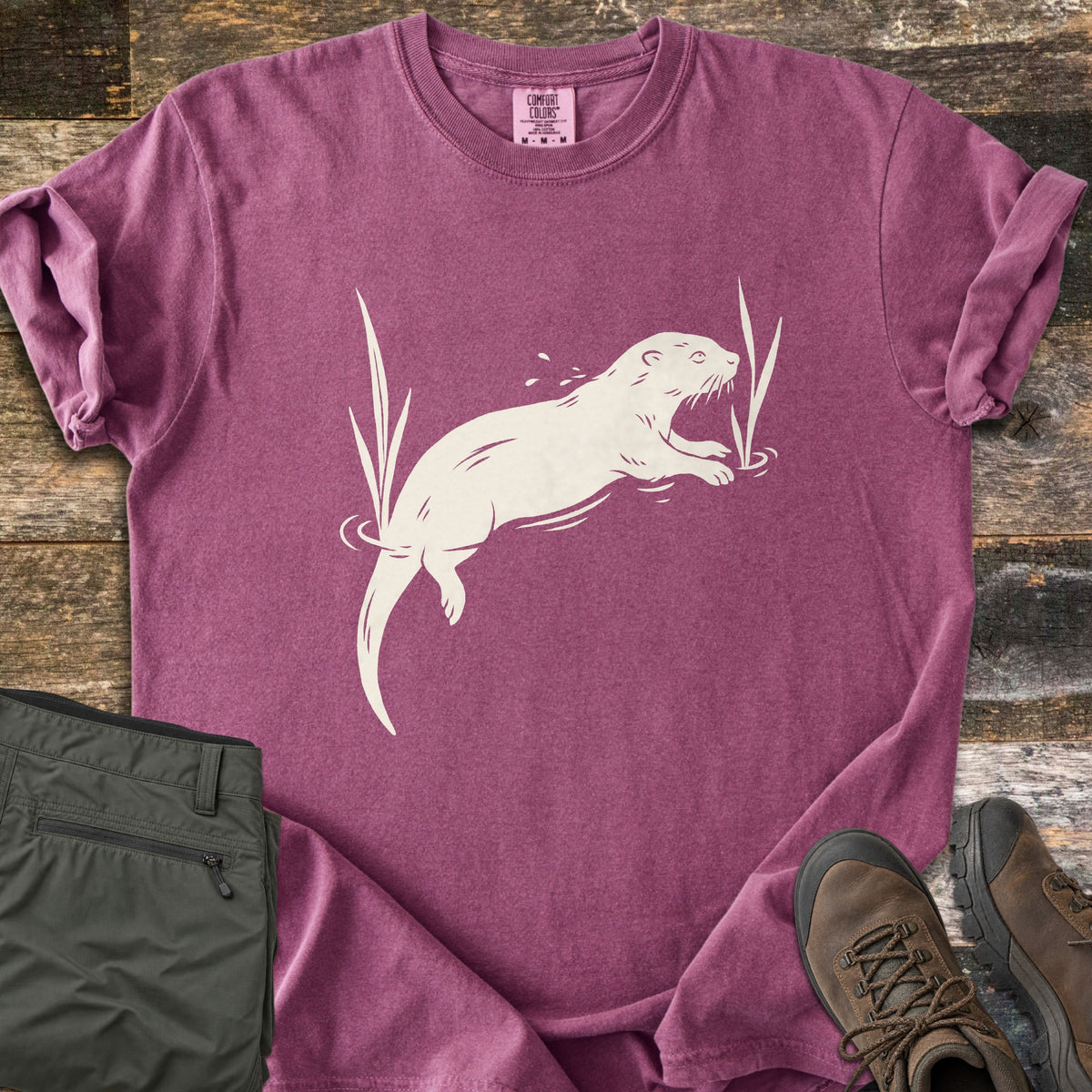 Swimming Otter Comfort Colors T-shirt in Berry, laid flat. A cream print of an otter swimming between tall reeds, with ripples and splashing droplets. Sleeves are rolled; olive hiking pants and brown hiking boots sit on weathered wood planks.