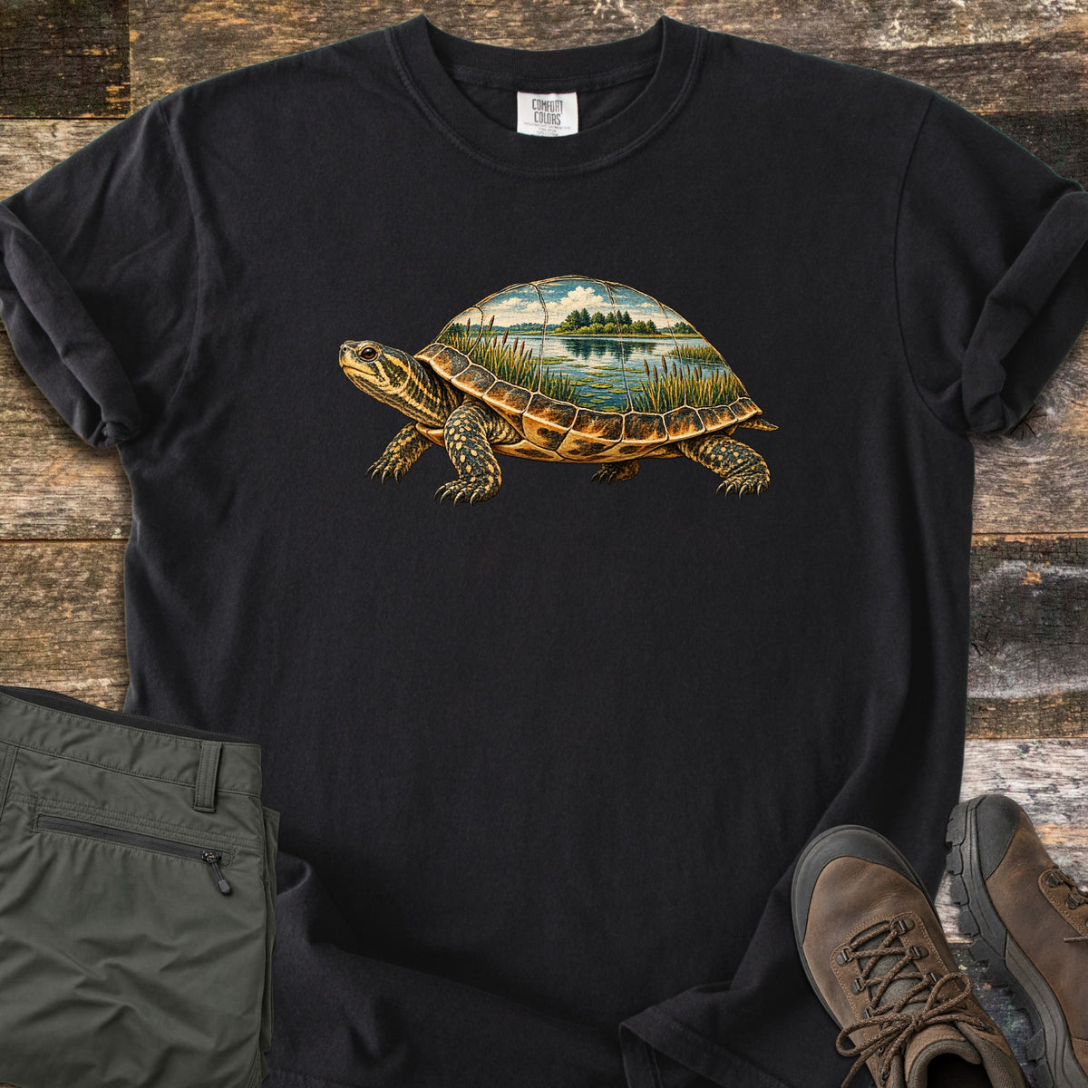 Turtle Pond Comfort Colors T-shirt in Black, laid flat. A turtle drawn in vintage illustration style, its shell showing a pond scene with cattails, water, trees and clouds. No words are printed. Olive hiking pants and brown hiking boots lie beside it on weathered wood.