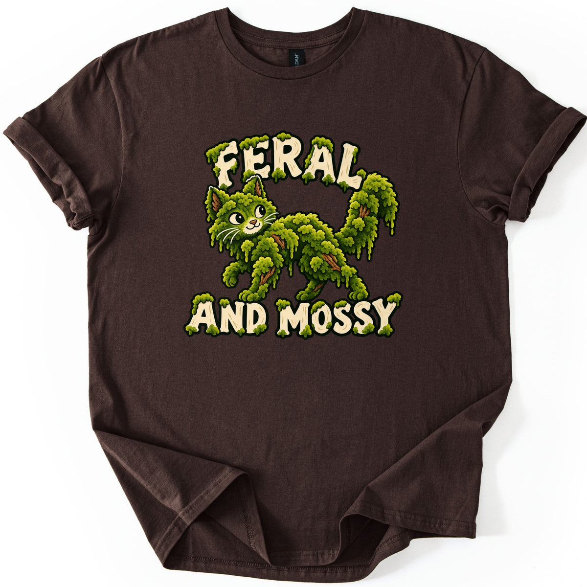 Feral and Mossy Comfort Colors T-shirt in Dark Chocolate, laid flat. A smiling cartoon cat made of dripping green moss with brown twigs showing through, between moss-covered cream letters reading Feral and Mossy. The shirt lies on white with cuffed sleeves and a folded hem.