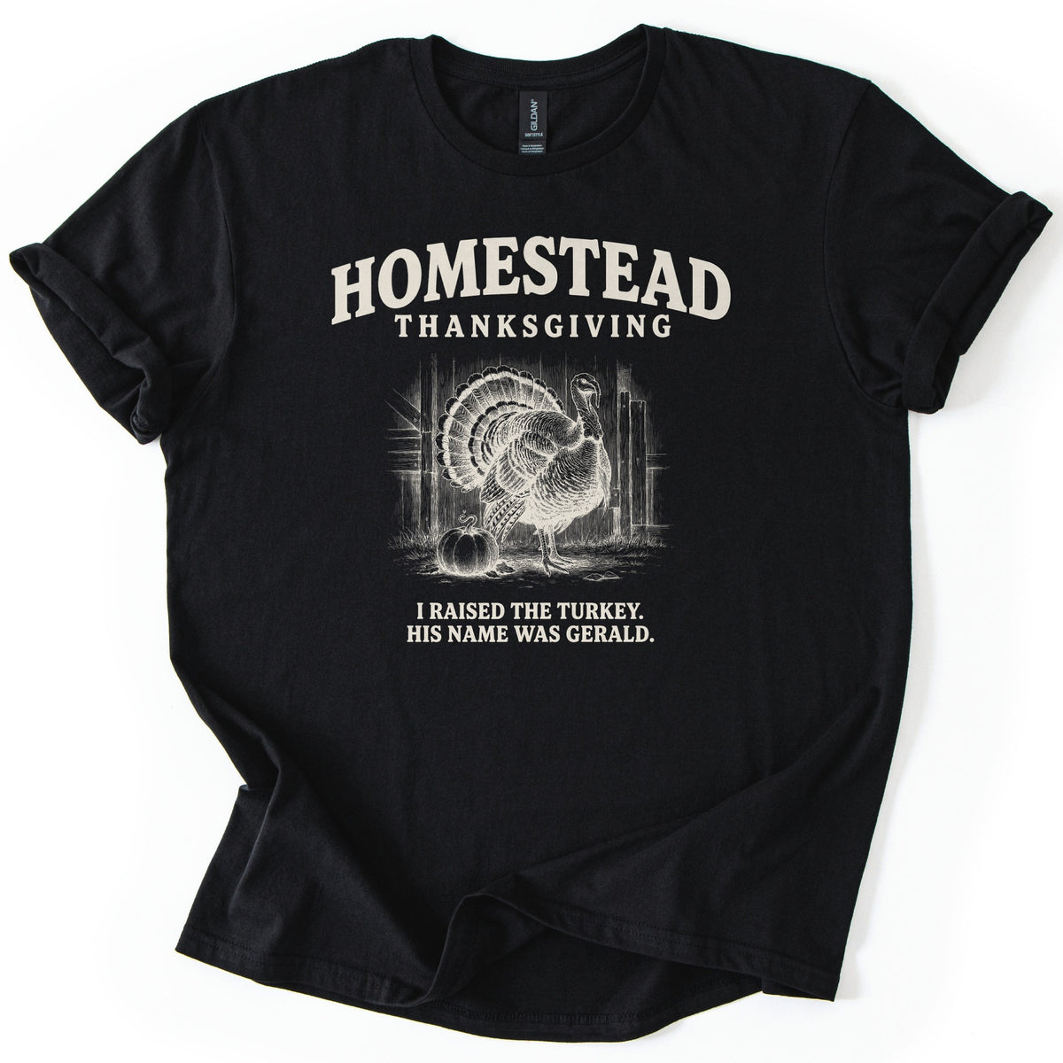 Homestead Thanksgiving Comfort Colors T-shirt in Black, laid flat. Cream sketch of a fanned-tail turkey beside a pumpkin by a wooden barn, under arched Homestead Thanksgiving and above I Raised the Turkey. His Name Was Gerald. Rolled sleeves, white background.