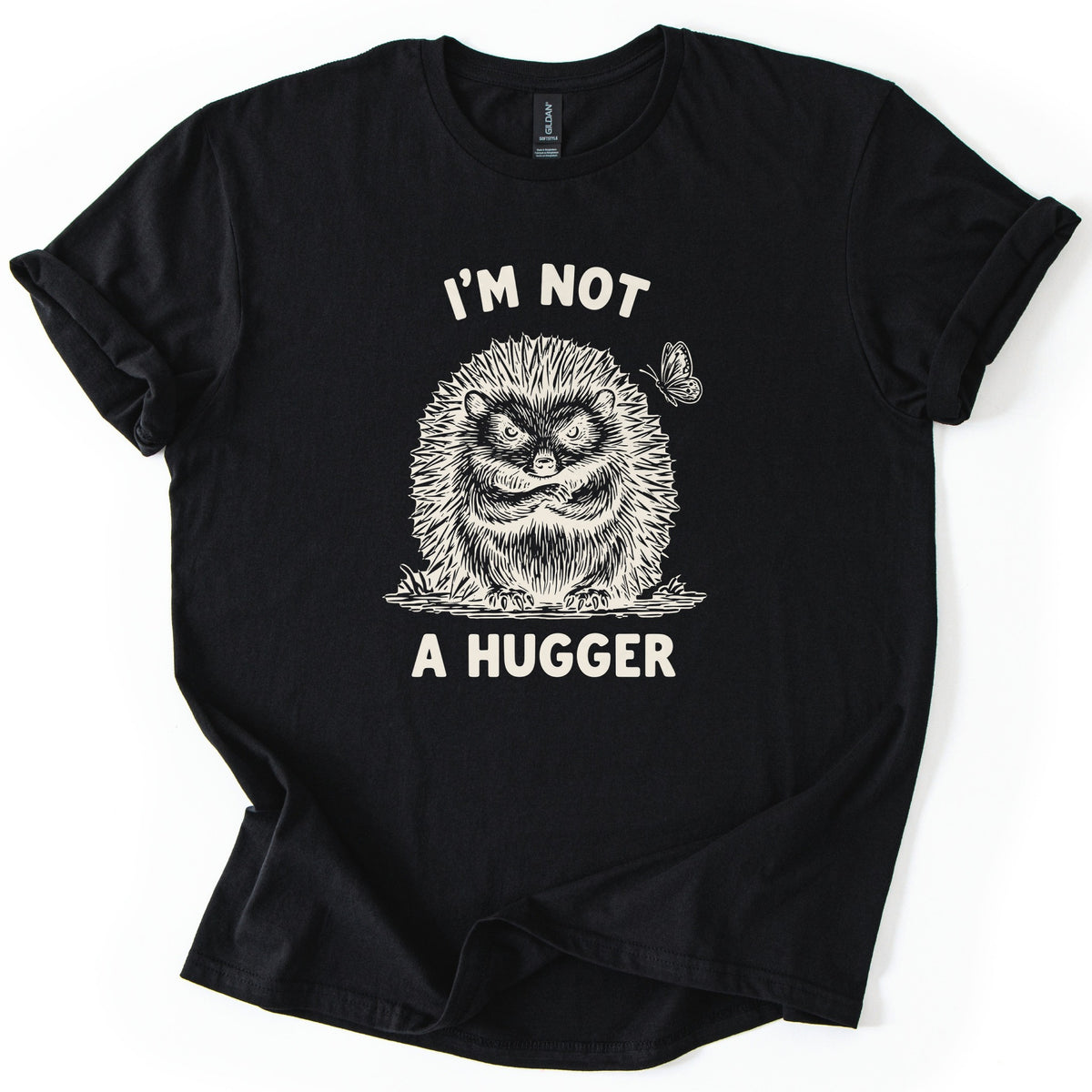 I'm Not A Hugger Comfort Colors T-shirt in Black, laid flat. A scowling sketch-style hedgehog with crossed arms, a butterfly beside it, sits between the words I'm Not A Hugger in cream. The sleeves are rolled on a white background.