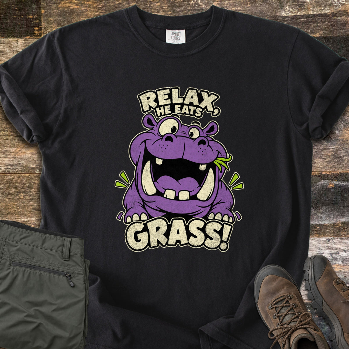 Relax He Eats Grass Comfort Colors T-shirt in Black, laid flat. A grinning purple cartoon hippo with a sprig of green grass in its mouth sits between the words Relax, He Eats and Grass! Folded olive pants and brown hiking boots lie beside it on weathered wood planks.