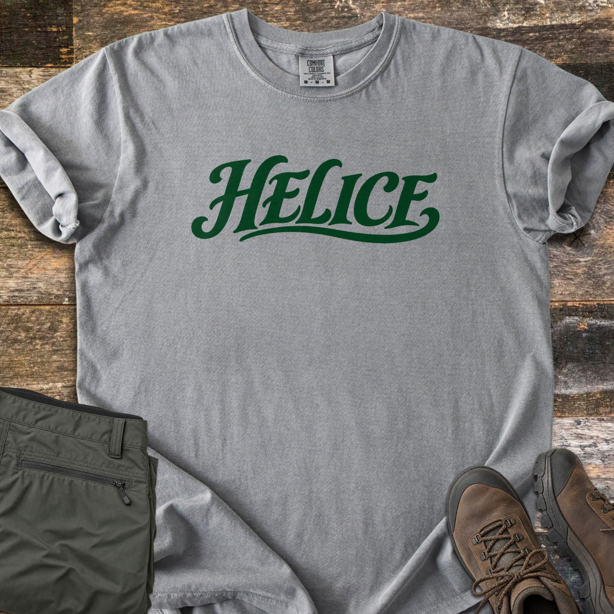 Helice Comfort Colors T-shirt in Grey, laid flat. The word Helice is printed in dark green script lettering with a swash underline across the chest. Sleeves are rolled, on a rustic wood floor beside folded olive cargo pants and brown leather hiking boots.