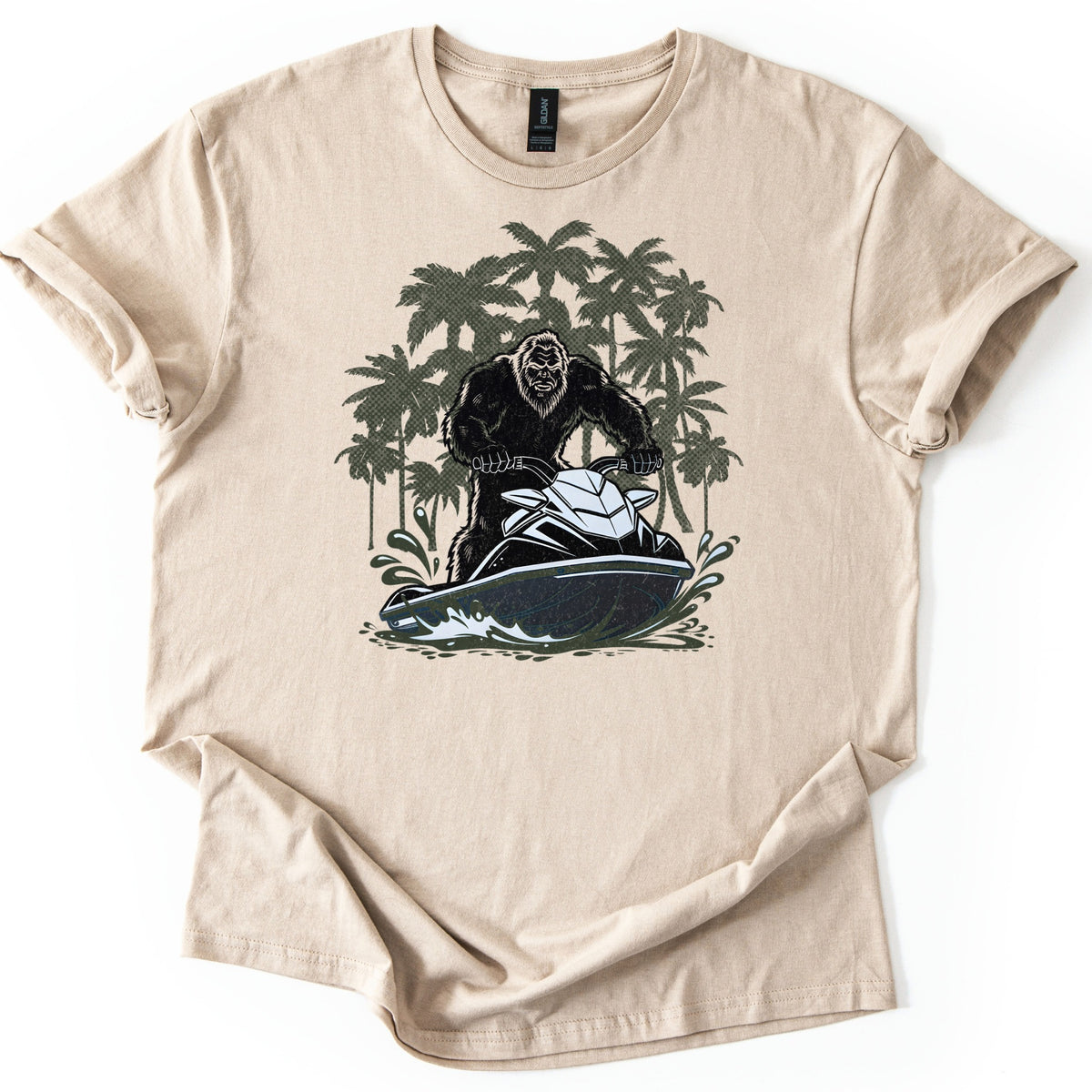 Jet Ski Squatch Comfort Colors T-shirt in Sand, laid flat. A black-furred Bigfoot rides a black and white jet ski through splashing water, with olive green palm trees behind him. No words are printed. The shirt lies on a white background with rolled sleeves.