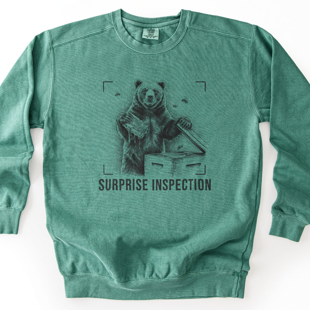 Surprise Inspection Comfort Colors crewneck sweatshirt in Light Green, laid flat. A sketched bear holds a dripping honeycomb and lifts the lid of a wooden beehive as bees fly nearby, inside camera viewfinder corners above the words SURPRISE INSPECTION in dark ink. Laid on a white surface.