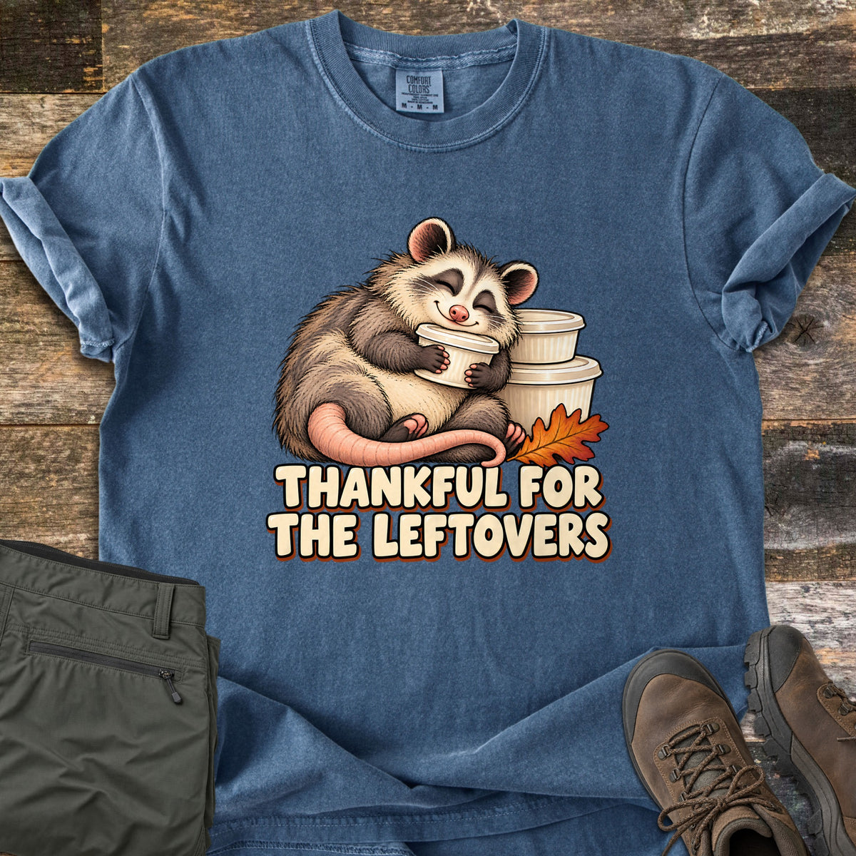 Thankful For The Leftovers Comfort Colors T-shirt in Denim, laid flat. A smiling opossum hugs a stack of white food containers beside an orange oak leaf above the words Thankful For The Leftovers. The shirt lies on dark wood with olive pants and brown hiking boots.