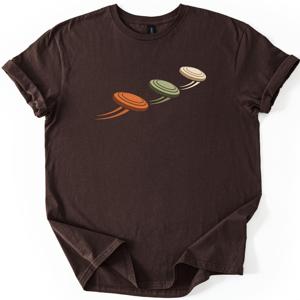 Flying Clays Comfort Colors T-shirt in Dark Chocolate, laid flat. Three clay targets in orange, olive green and cream fly upward in a diagonal line across the chest, each trailing motion streaks, with no words. The shirt lies on a white surface with cuffed sleeves.