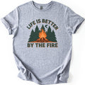 Life Is Better By The Fire Comfort Colors T-shirt in Sport Grey, laid flat. A campfire of crossed logs burns in front of dark green pine trees under a crescent moon, with LIFE IS BETTER arched above and BY THE FIRE below in dark green letters. Cuffed sleeves, white background.