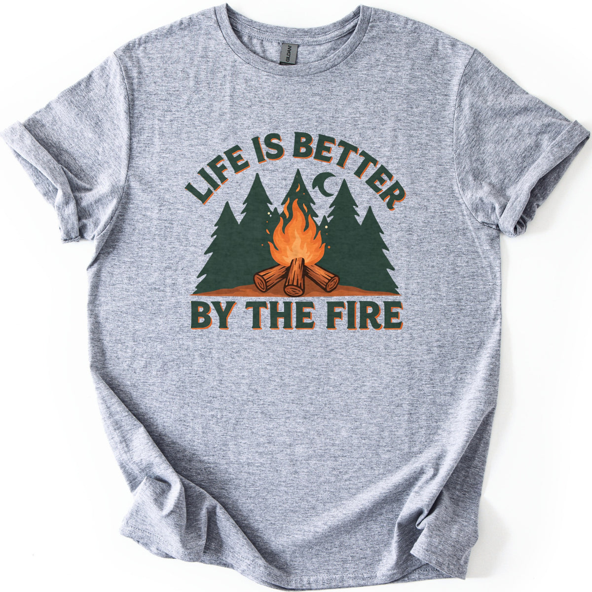 Life Is Better By The Fire Comfort Colors T-shirt in Sport Grey, laid flat. A campfire of crossed logs burns in front of dark green pine trees under a crescent moon, with LIFE IS BETTER arched above and BY THE FIRE below in dark green letters. Cuffed sleeves, white background.