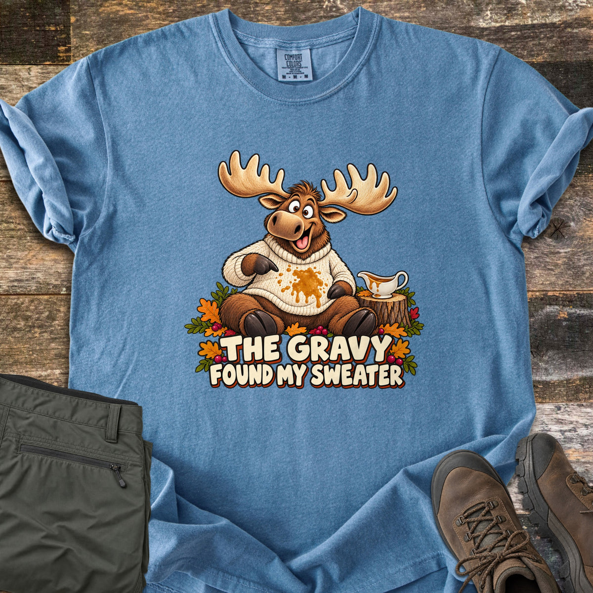 The Gravy Found My Sweater Comfort Colors T-shirt in Blue Jean, laid flat. A cartoon moose in a cream knit sweater points at a gravy stain beside a gravy boat on a stump, above the words The Gravy Found My Sweater. Olive pants and brown boots sit on a wood floor.