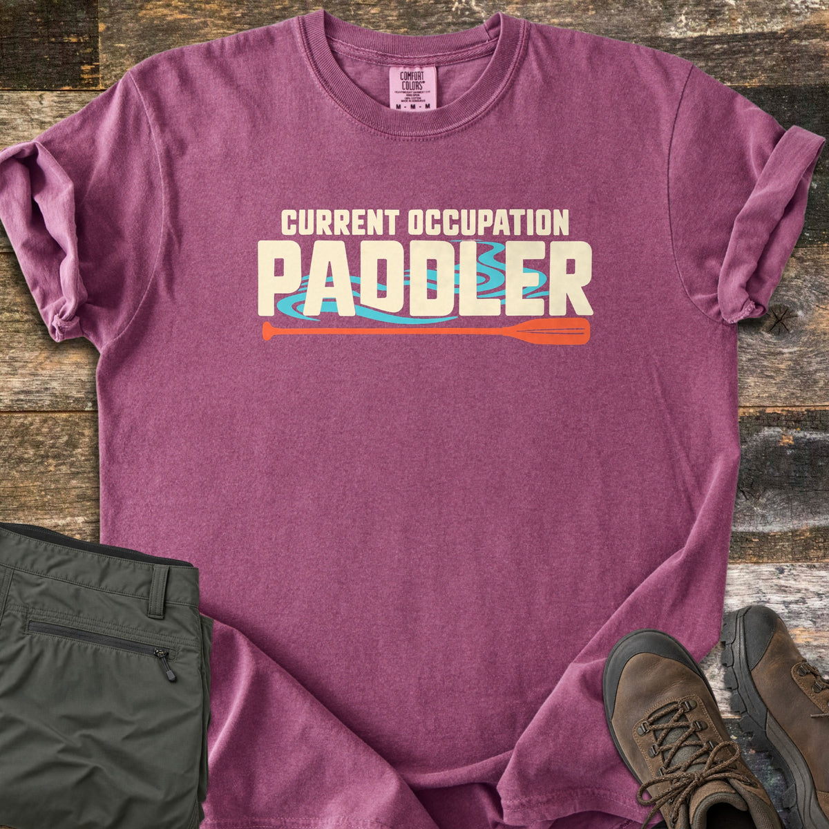 Current Occupation Paddler Comfort Colors T-shirt in Berry, laid flat. Cream block letters read Current Occupation Paddler over turquoise water swirls, with an orange canoe paddle beneath. Sleeves are rolled on a weathered wood surface beside olive hiking pants and brown hiking boots.