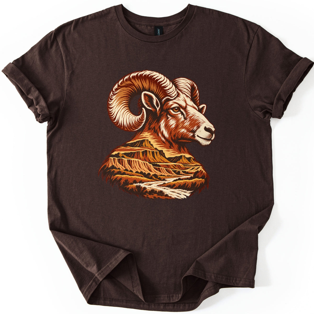 Bighorn Badlands Comfort Colors T-shirt in Dark Chocolate, laid flat. A bighorn ram's head with large curled horns, drawn in rust, orange and cream, blends into badlands buttes and a winding river at its neck. No words are printed. The shirt lies on a white surface with rolled sleeves.