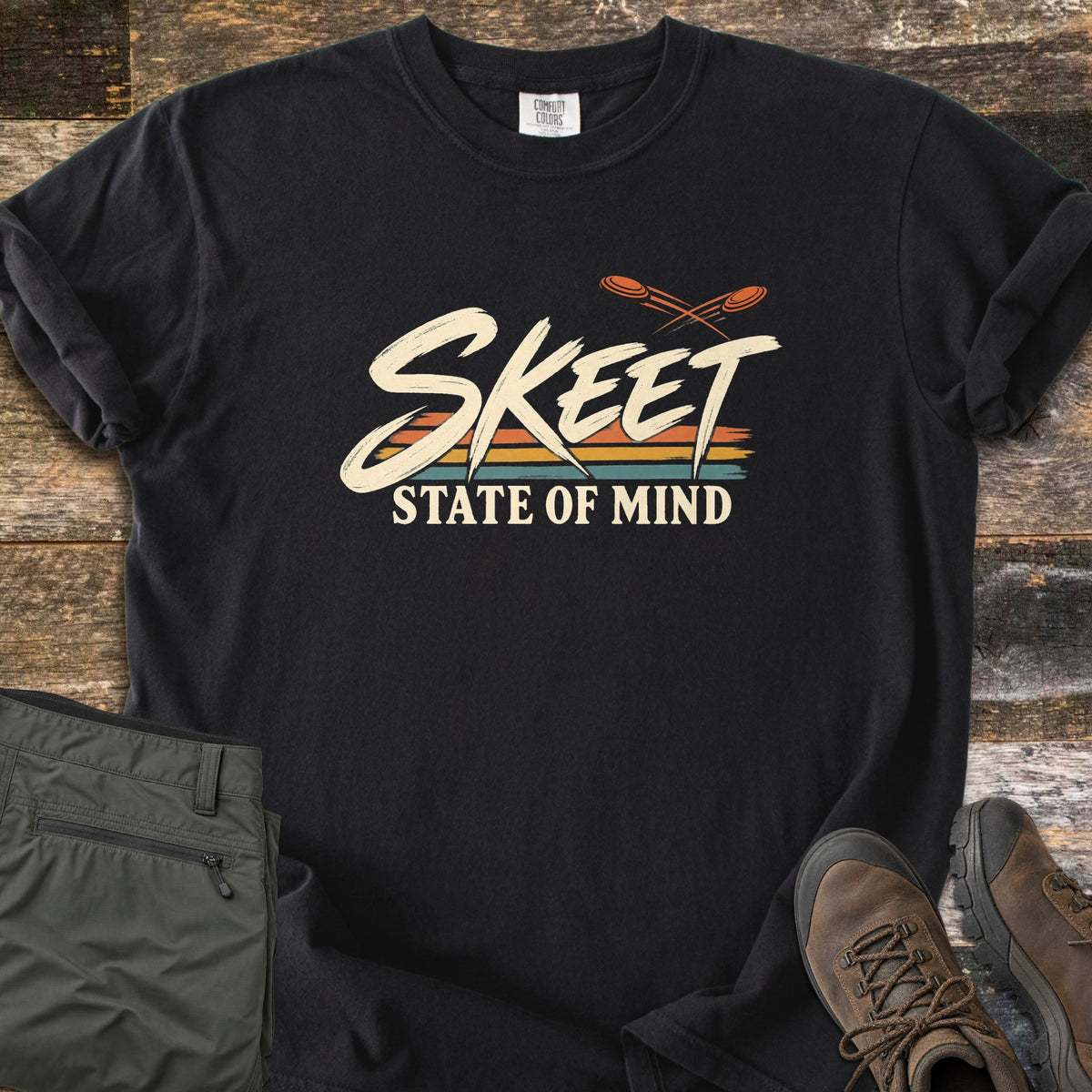Skeet State of Mind Comfort Colors T-shirt in Black, laid flat. Brushy cream letters spell Skeet over orange, yellow and teal stripes, with two orange clay targets flying above and State of Mind below. Olive pants and brown hiking boots sit on rustic wood planks.