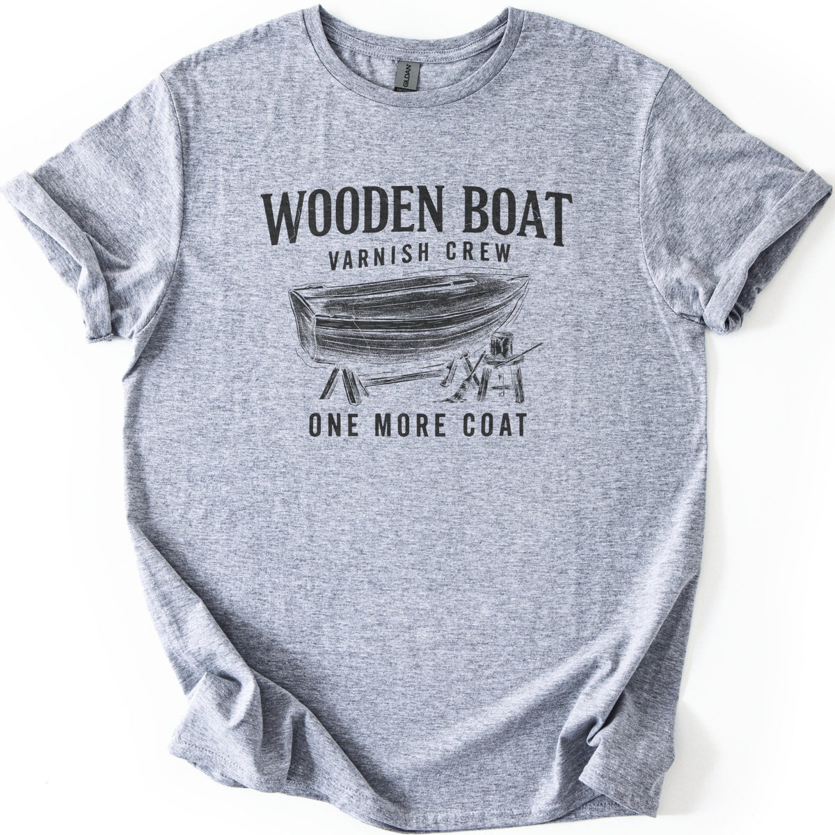 Wooden Boat Varnish Crew Comfort Colors T-shirt in Sport Grey, laid flat. Black print reads Wooden Boat, Varnish Crew above a sketch of a wooden rowboat on sawhorses beside a stool with a varnish can and brush, then One More Coat. Sleeves cuffed on a white background.