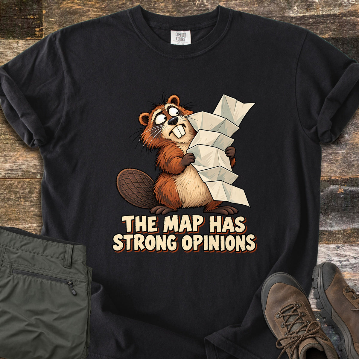 The Map Has Strong Opinions Comfort Colors T-shirt in Black, laid flat. A cartoon beaver with wide, crossed eyes clutches a tall, unfolding paper map above the words The Map Has Strong Opinions. Sleeves are rolled, beside green hiking pants and brown hiking boots on weathered wood.