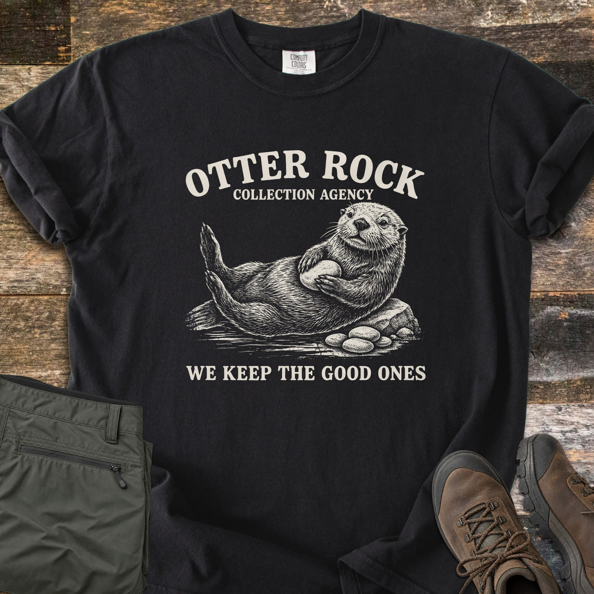 Otter Rock Collection Agency Comfort Colors T-shirt in Black, laid flat. A cream sketch of a sea otter lying on a rock holding a stone, under the words Otter Rock Collection Agency, with We Keep the Good Ones below. Shown on wood planks with olive pants and brown hiking boots.