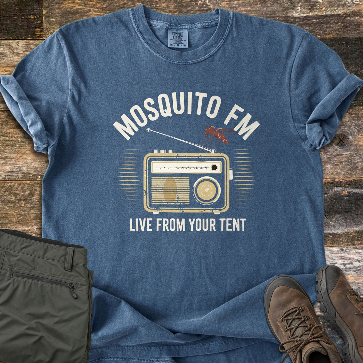 Mosquito FM Comfort Colors T-shirt in Denim, laid flat. A vintage radio with a mosquito perched on its antenna sits under the words Mosquito FM and above Live From Your Tent. Olive hiking pants and brown hiking boots rest beside it on weathered wood planks.