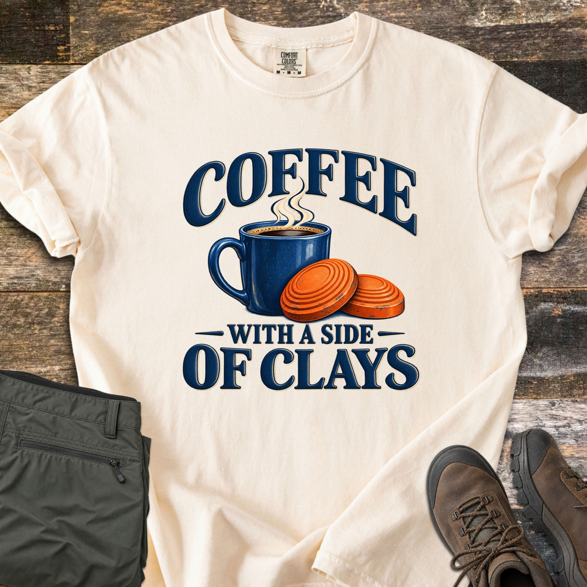 Coffee with a Side of Clays Comfort Colors T-shirt in Ivory, laid flat. A steaming blue mug of coffee beside two orange clay targets, with the words Coffee with a Side of Clays in navy. Sleeves rolled, on rustic wood with folded green pants and brown hiking boots.