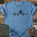 Bigfoot Heartbeat Comfort Colors T-shirt in Blue Jean, laid flat. A black heartbeat line runs across the chest with a walking Bigfoot silhouette in the middle; there are no words. The shirt lies on weathered wood beside folded olive hiking pants and brown hiking boots.