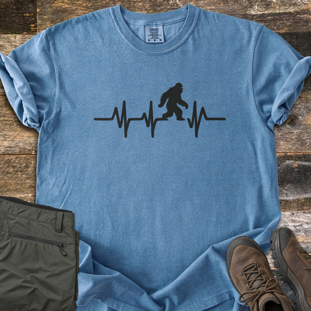 Bigfoot Heartbeat Comfort Colors T-shirt in Blue Jean, laid flat. A black heartbeat line runs across the chest with a walking Bigfoot silhouette in the middle; there are no words. The shirt lies on weathered wood beside folded olive hiking pants and brown hiking boots.