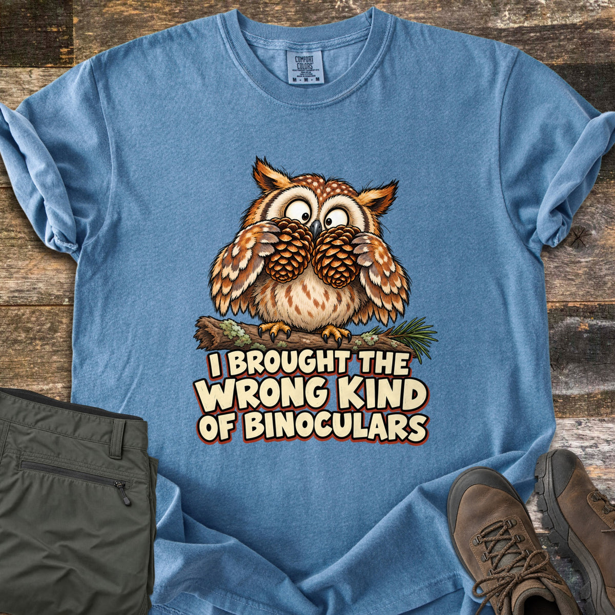 I Brought The Wrong Kind Of Binoculars Comfort Colors T-shirt in Blue Jean, laid flat. A wide-eyed cartoon owl on a mossy branch holds two pinecones to its eyes like binoculars above the words I Brought The Wrong Kind Of Binoculars. Olive hiking pants and brown hiking boots lie beside it on weathered wood.