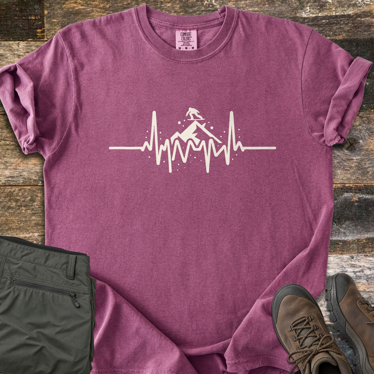 Snow Boarder Heartbeat Comfort Colors T-shirt in Berry, laid flat. A cream heartbeat line rises into a mountain peak with a snowboarder riding on top, surrounded by snow dots. Folded olive hiking pants and brown leather boots sit beside it on weathered wood planks.